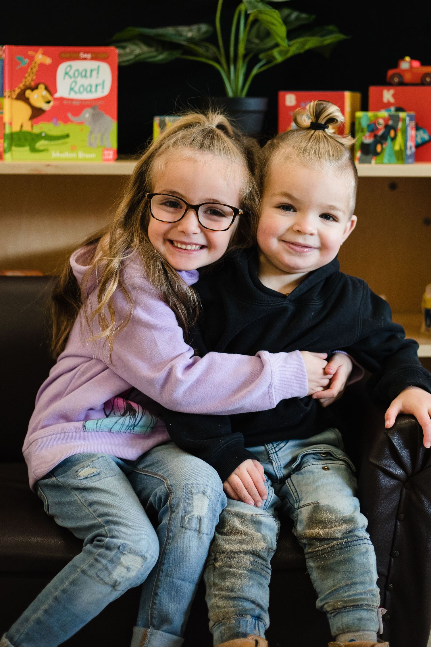 Two children sit together on a dark couch, wearing casual clothes with a bookshelf visible in the background.