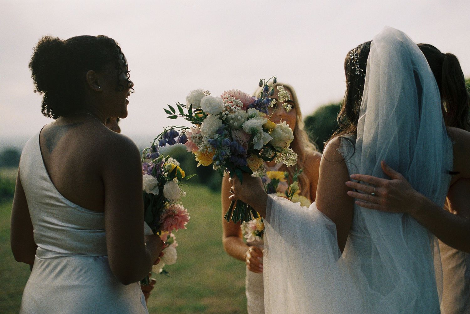 Bride in white gown and veil holding floral bouquet with bridesmaids outdoors at wedding celebration.