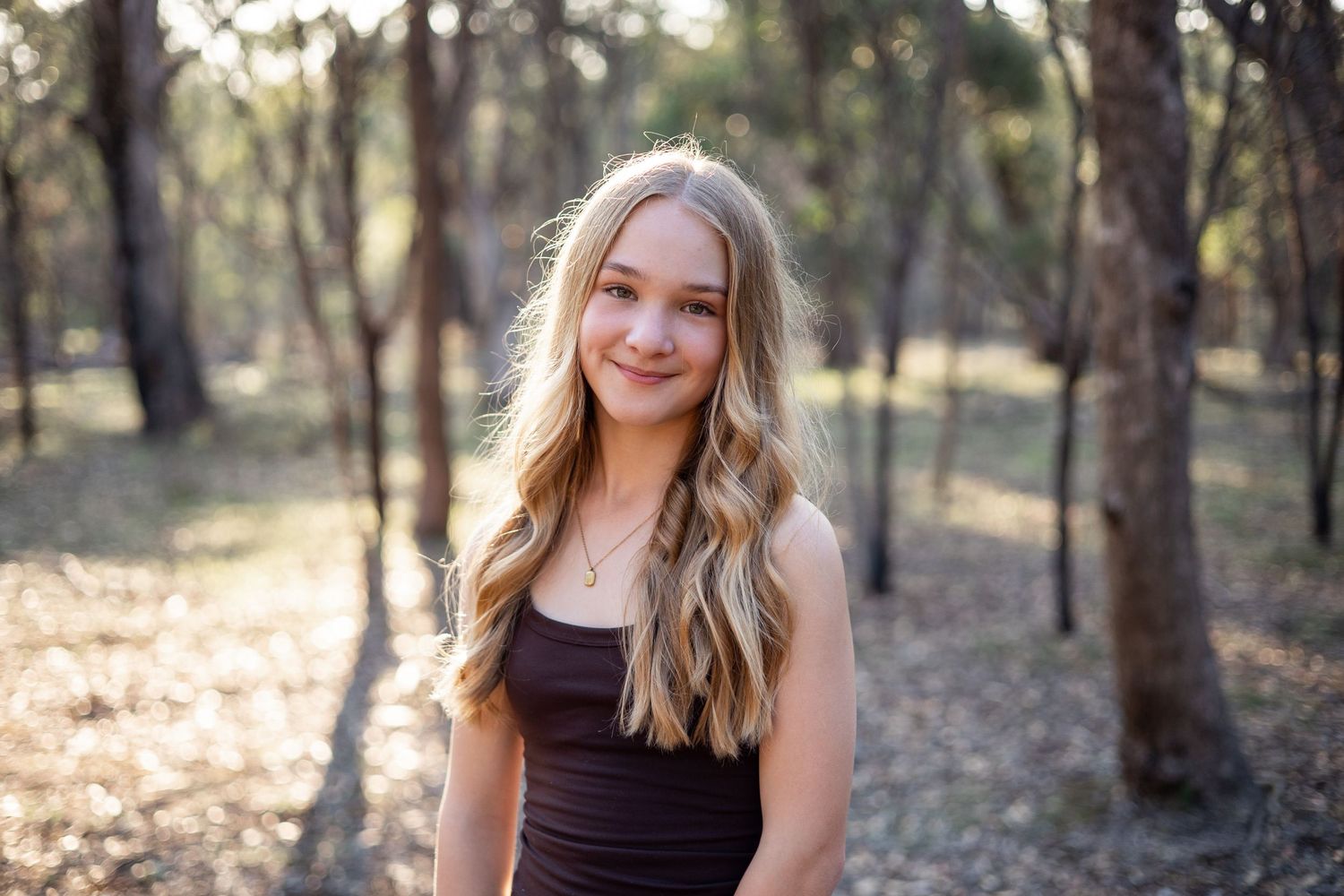 Young blonde woman smiling in a sunlit forest wearing a brown top, perfect for outdoor portrait photography.