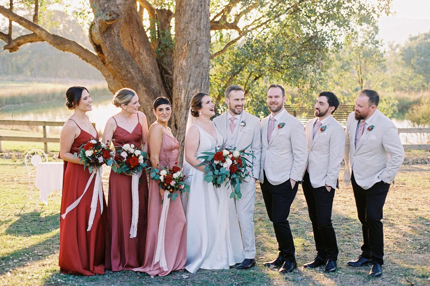 Wedding party poses outdoors near large tree, bridesmaids in red and pink gowns, groomsmen in beige suits holding bouquets.