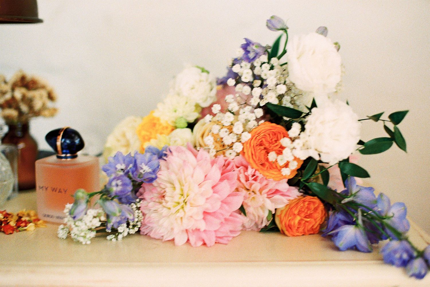 Colorful floral bouquet with pink dahlias, orange roses, white hydrangeas, and blue flowers on a white surface near a perfume bottle.