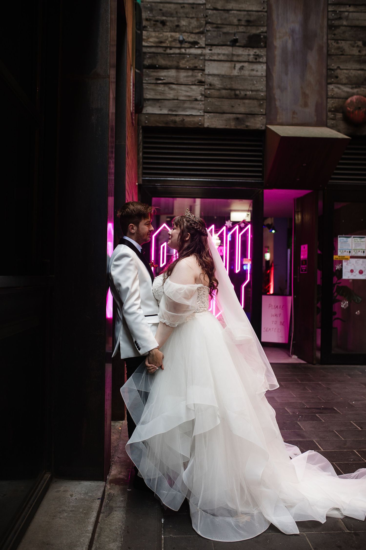 Bride and groom share a romantic kiss outside a venue with pink neon lights, bride wearing a flowing white tulle gown.