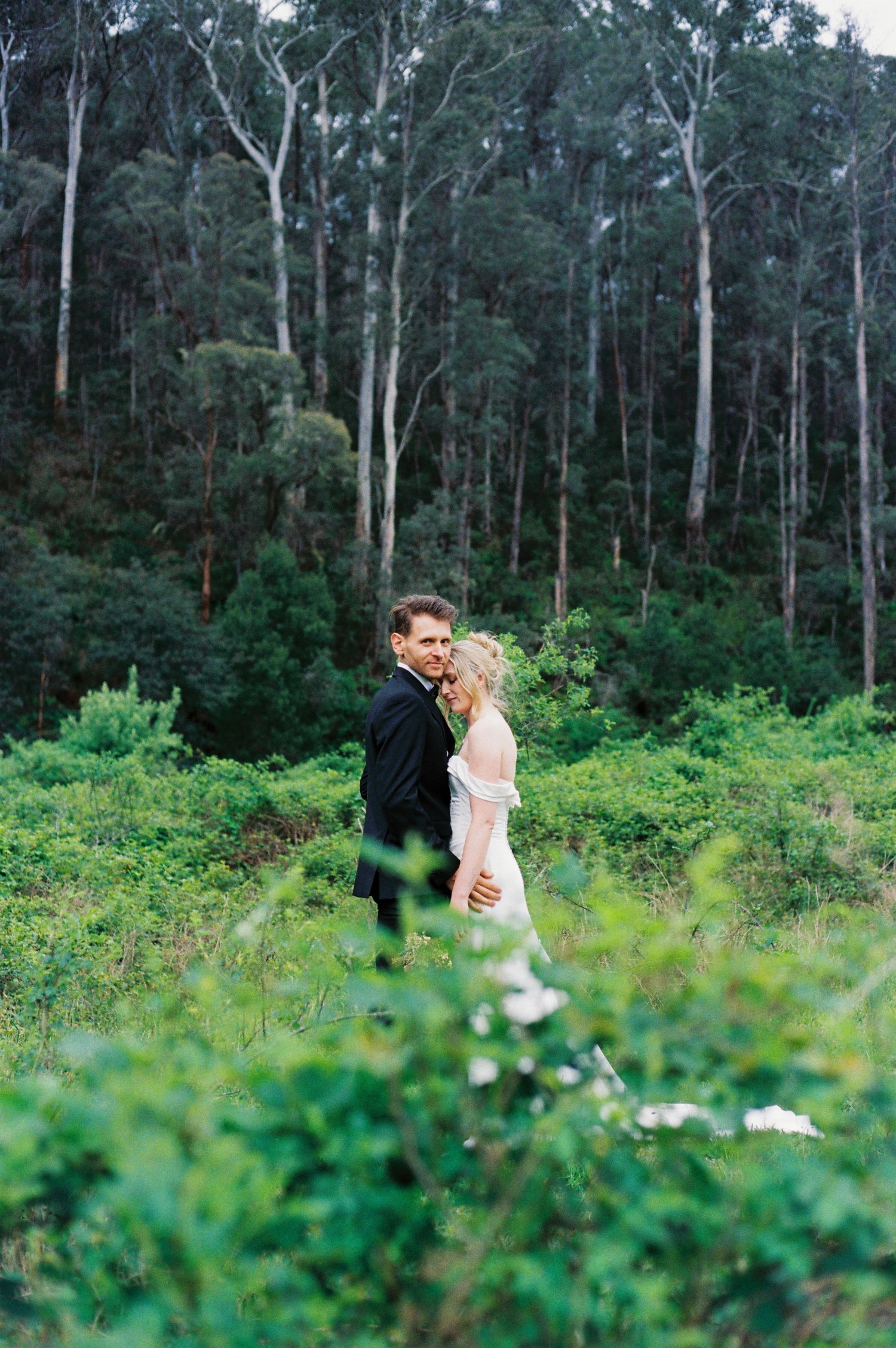 A couple stands together in a lush green forest with tall eucalyptus trees in the background.