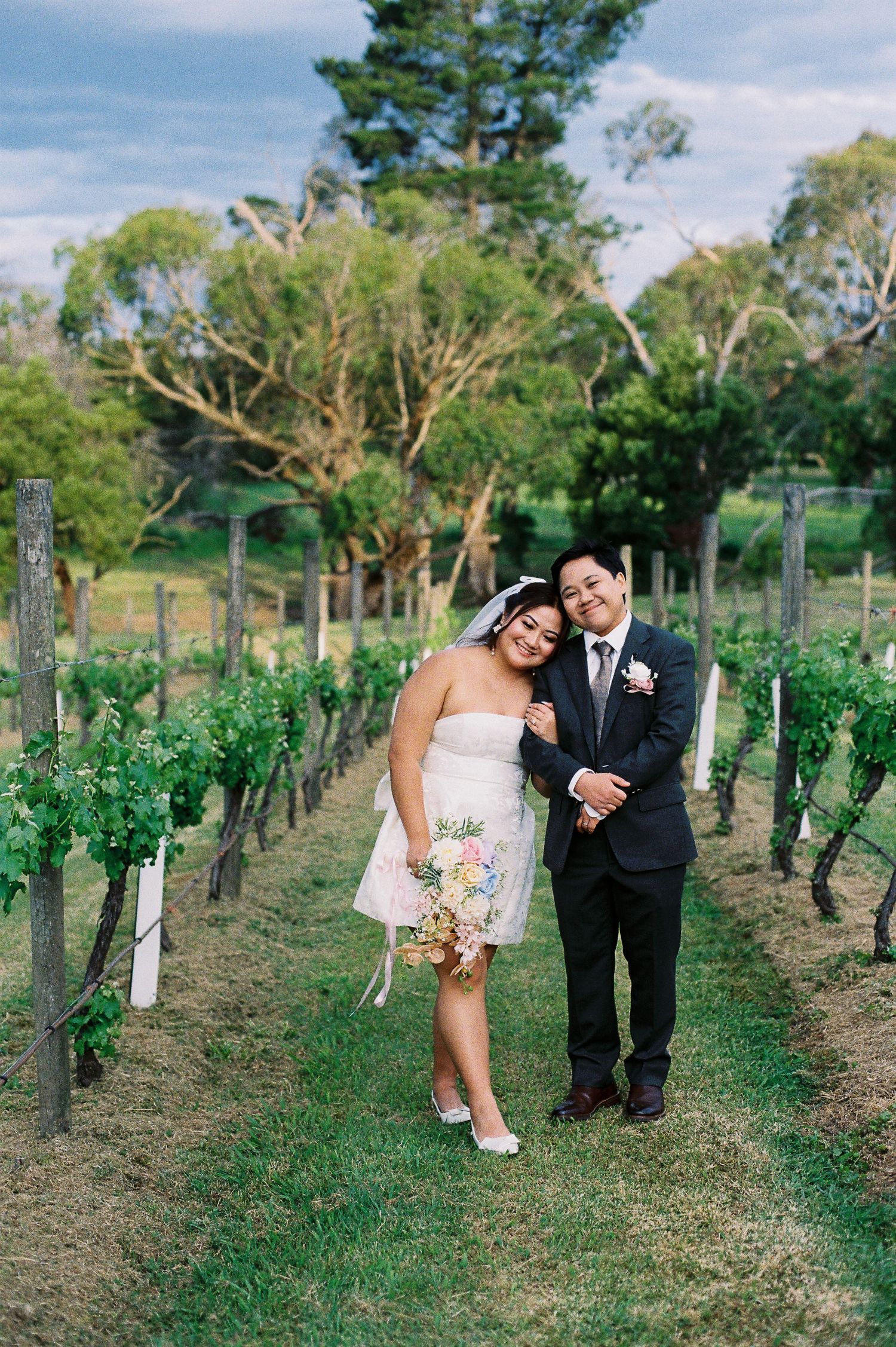 Bride and groom posing happily between vineyard rows, bride in short white dress holding bouquet, groom in dark suit.
