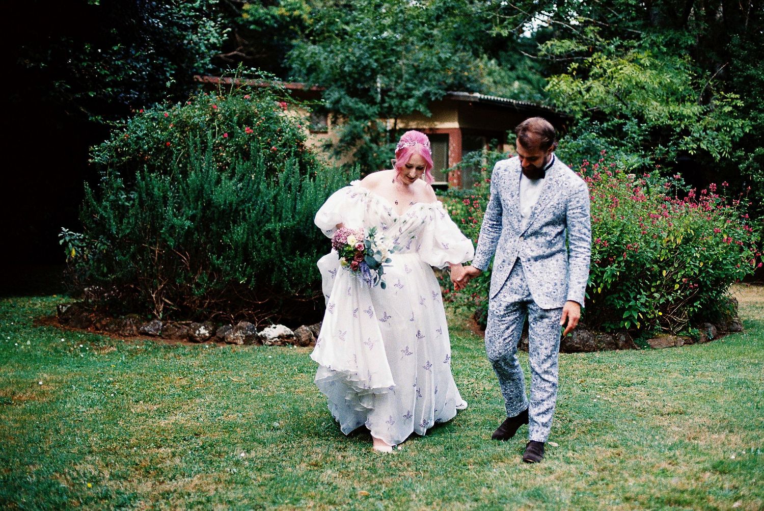 A couple in white and blue outfits dances playfully on a green lawn surrounded by lush garden landscaping.
