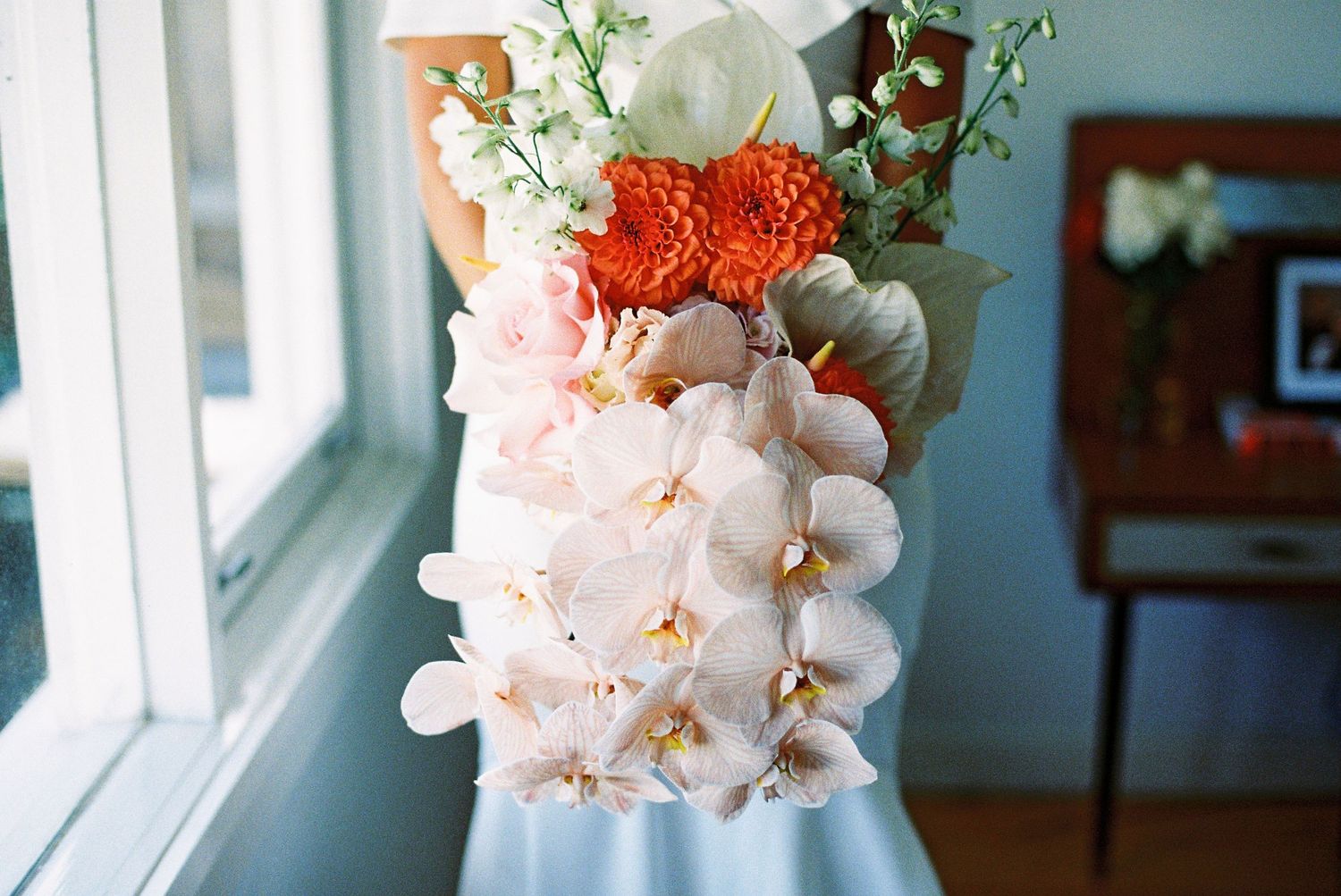 Elegant bridal bouquet featuring white orchids, orange chrysanthemums, and white ranunculus held near a sunlit window.