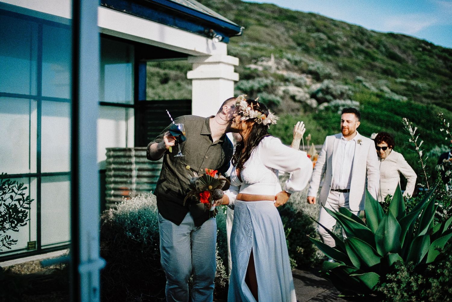 Couple shares a romantic kiss during outdoor wedding ceremony with guests watching against scenic hillside backdrop.