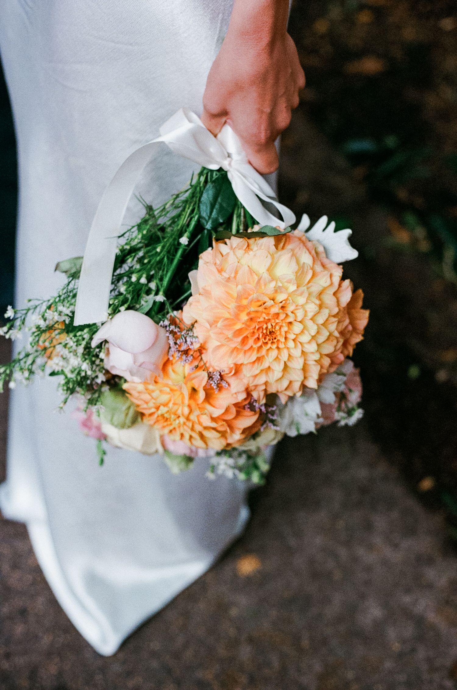 Bride holding a peach dahlia wedding bouquet with white ribbon, greenery, and mixed blooms in a bridal setting.