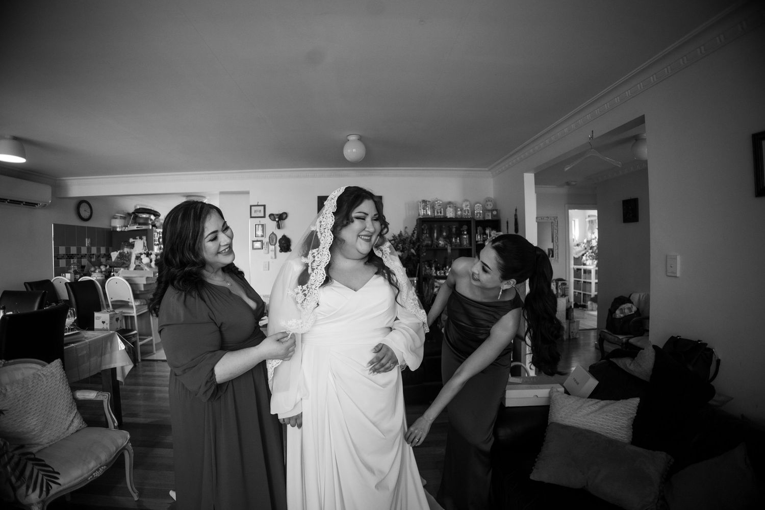 Black and white photo of a bride in a white gown being assisted by two bridesmaids in a cozy indoor setting.