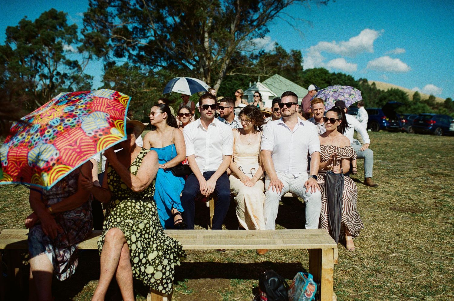 A group dressed in summer attire sits on a wooden bench with decorative parasols at an outdoor event on a sunny day.