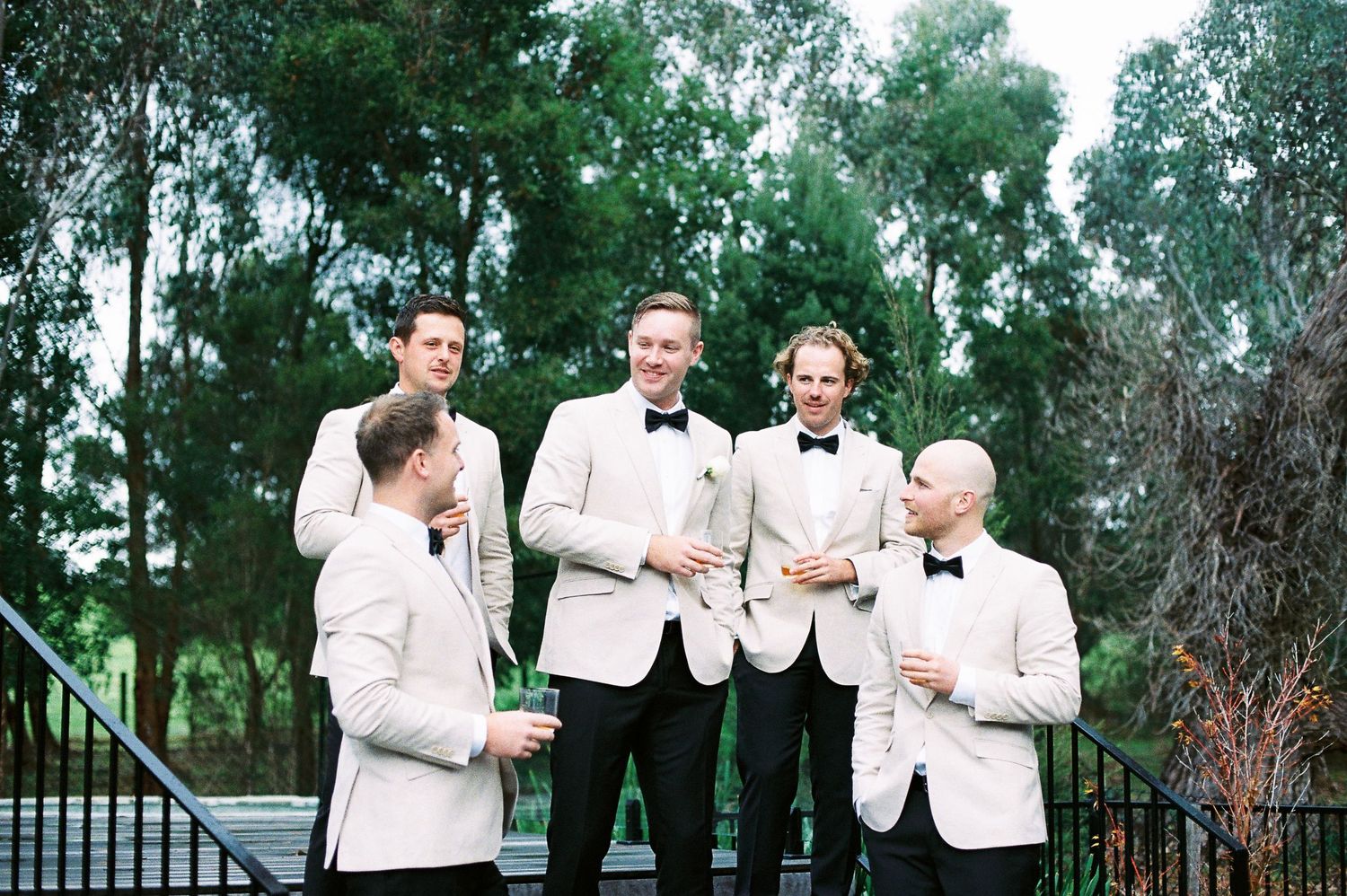 Five groomsmen in white tuxedo jackets and black bow ties laughing and celebrating outdoors at a wedding.
