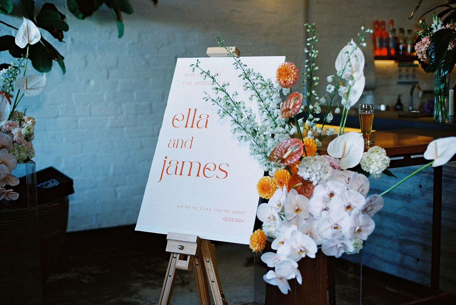 Wedding welcome sign for Ella and James on wooden easel with colorful floral arrangement of white and orange blooms.