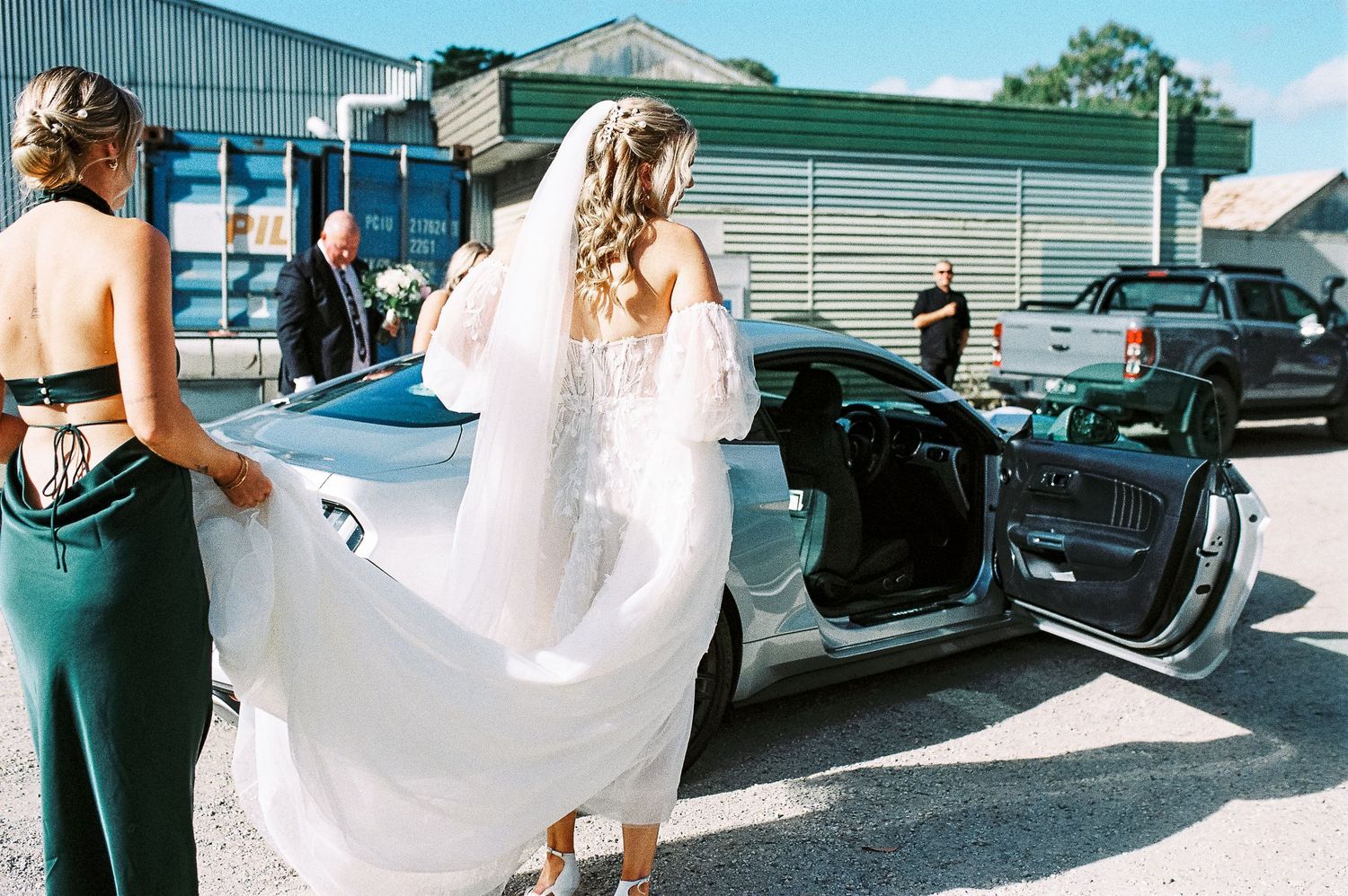 Bride in white lace gown with flowing veil stepping into a convertible car, assisted by bridesmaid in green dress.