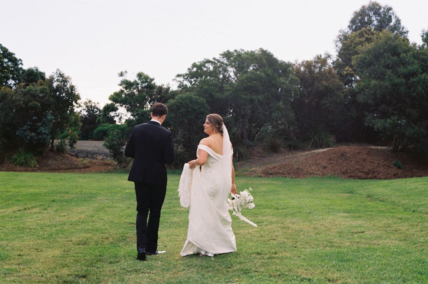 Bride and groom walking hand in hand across lush green lawn with trees in background at outdoor wedding.