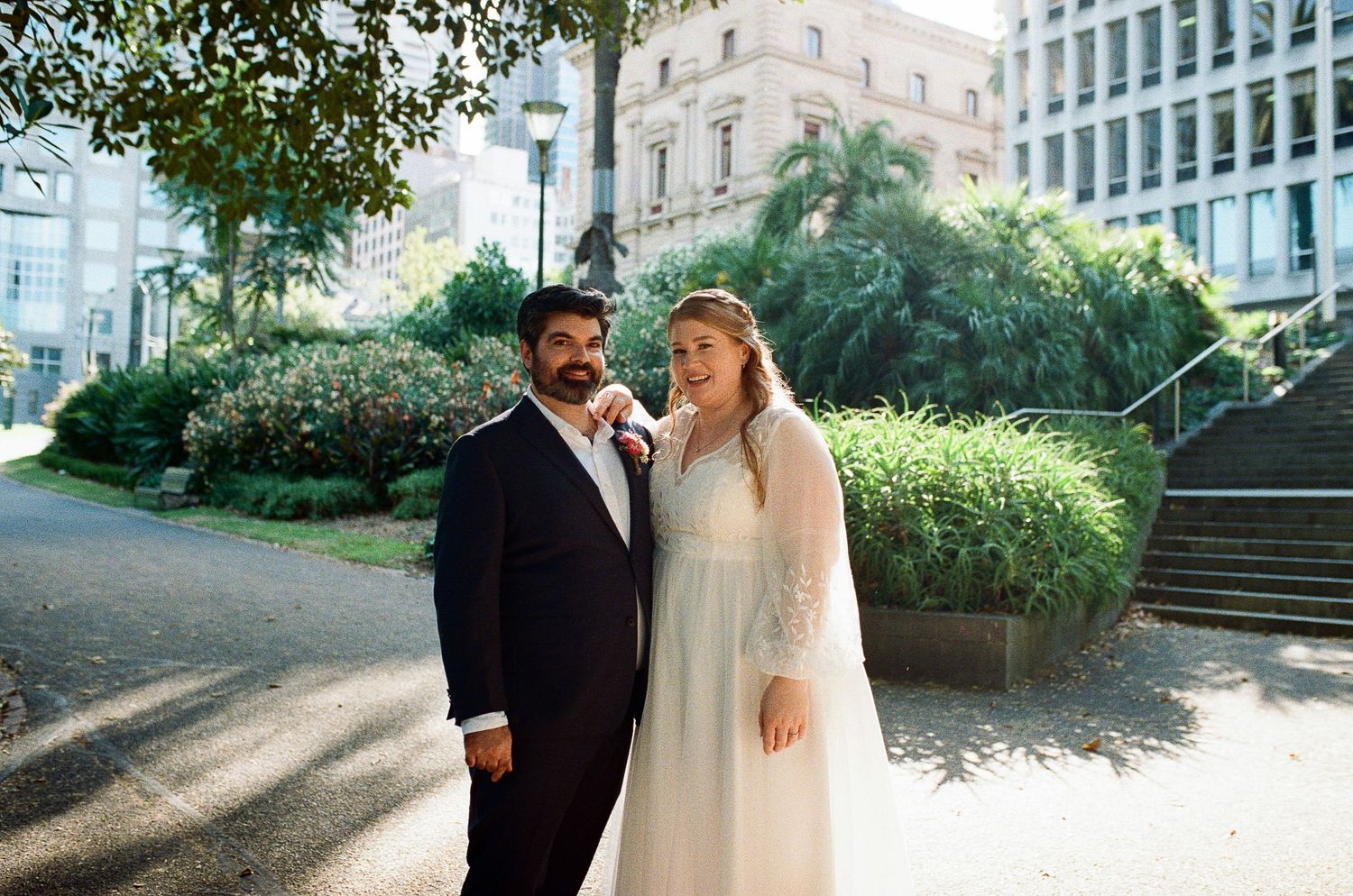 A couple in wedding attire shares an intimate moment in a garden setting with natural greenery and sunlight.