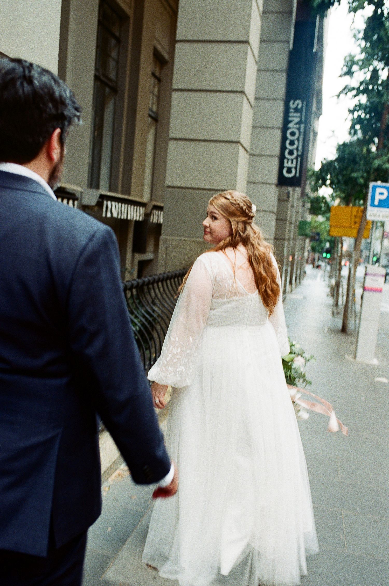 Bride in white lace gown walks with groom in navy suit on city sidewalk near Cecconi's restaurant.