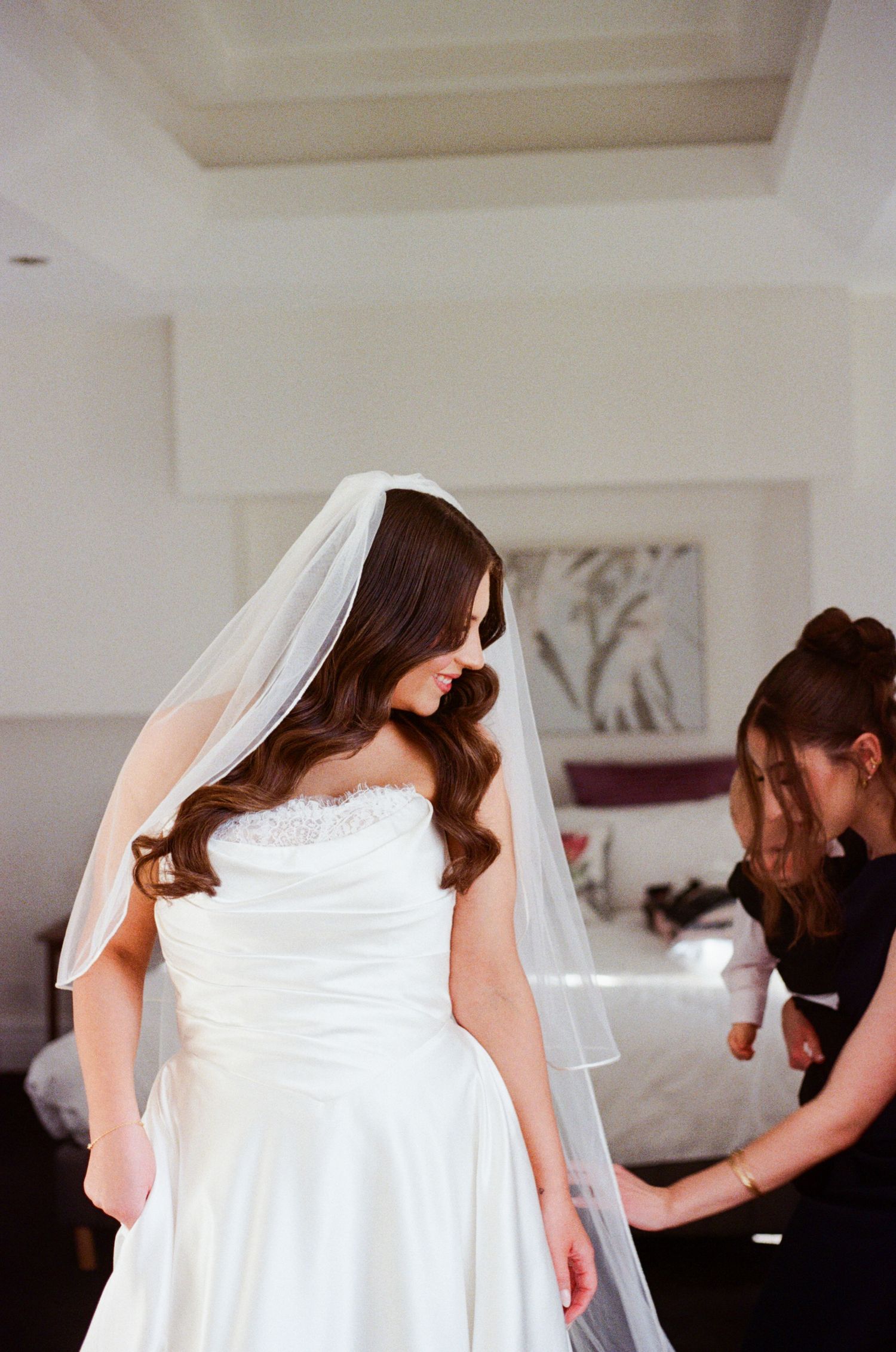 Bride in white strapless gown and veil with long brunette hair, assisted by bridesmaid in bright hotel room.