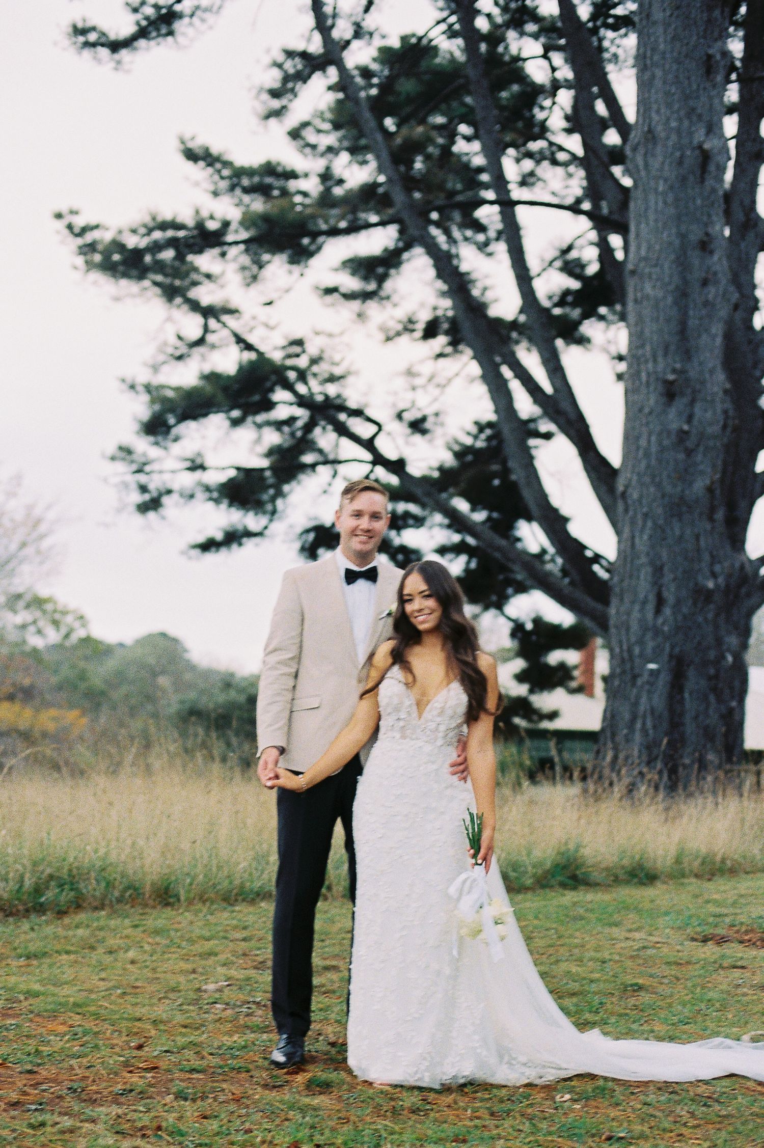 A smiling bride and groom pose outdoors in a field with a large tree, she in lace gown, he in tan suit with bow tie.
