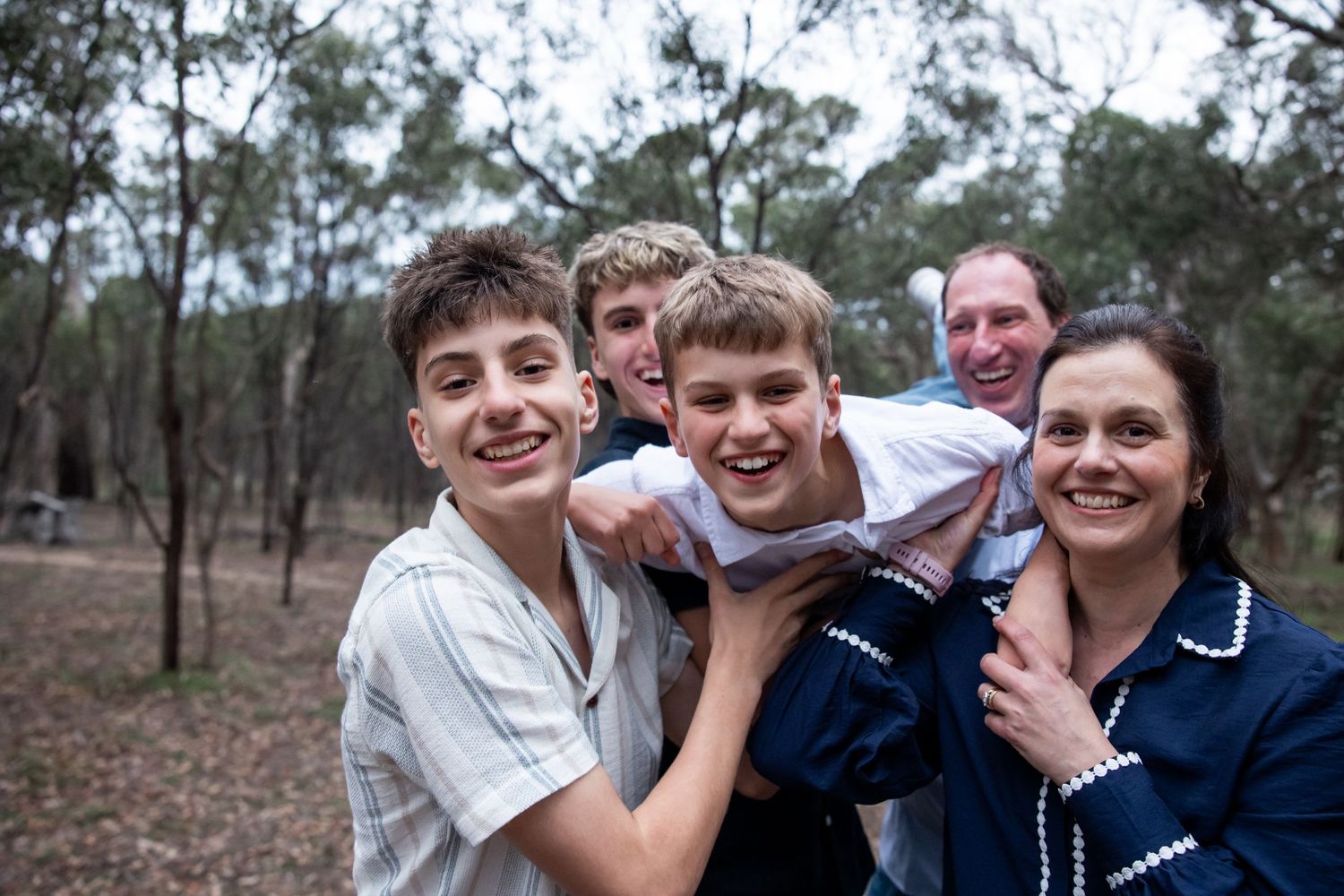 Happy family of four smiling outdoors in a bushland setting, with three teenage boys and a woman in a navy top.