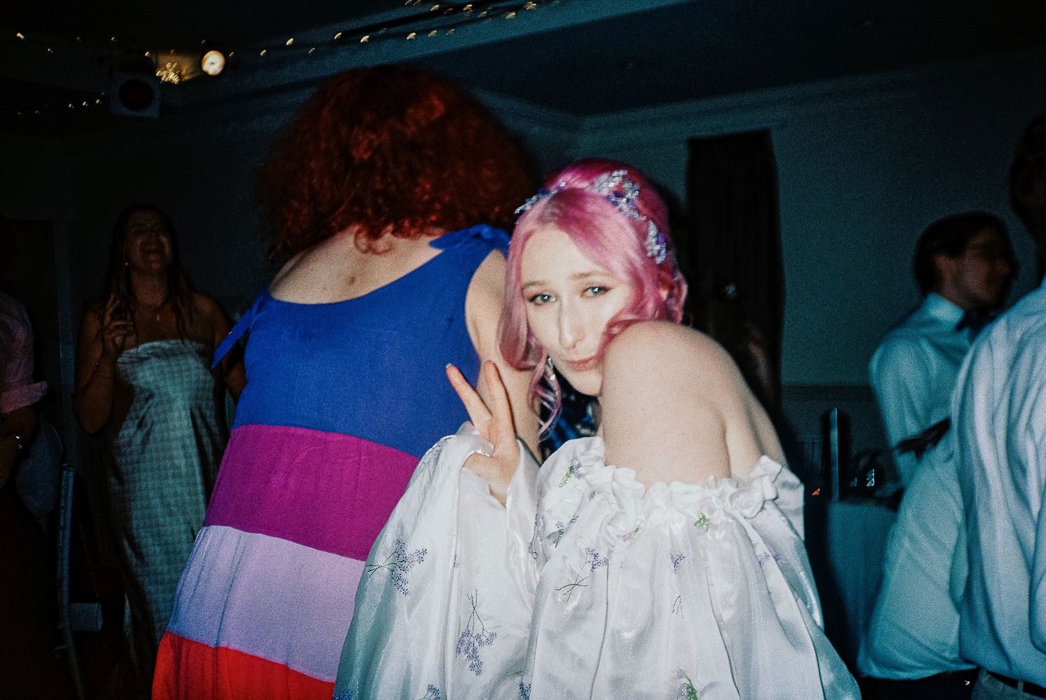 Two young women posing together at a party, one with red hair in colorful dress, other with pink hair in white off-shoulder top.
