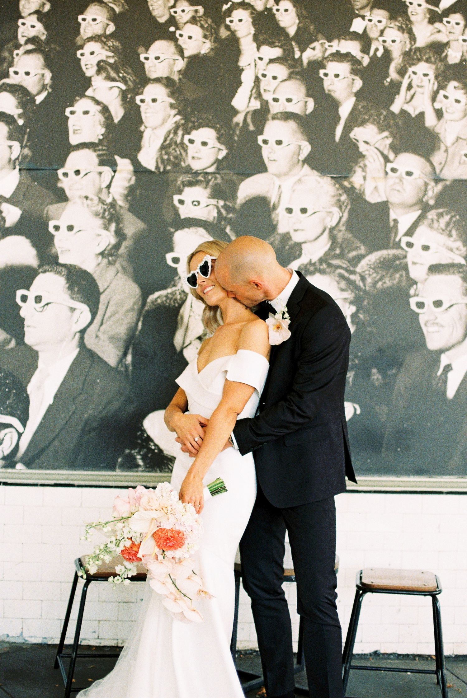 A couple sharing a romantic kiss in front of a vintage black and white photo backdrop showing a crowd wearing 3D glasses.