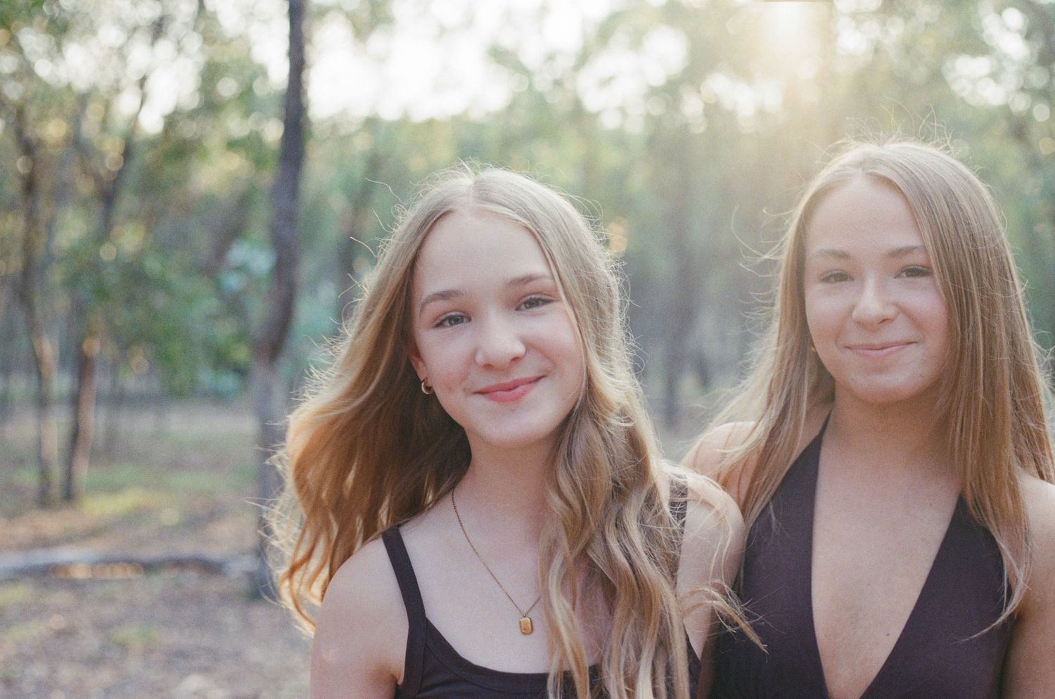 Two smiling teenage girls with long blonde hair posing outdoors in a sunlit forest setting.
