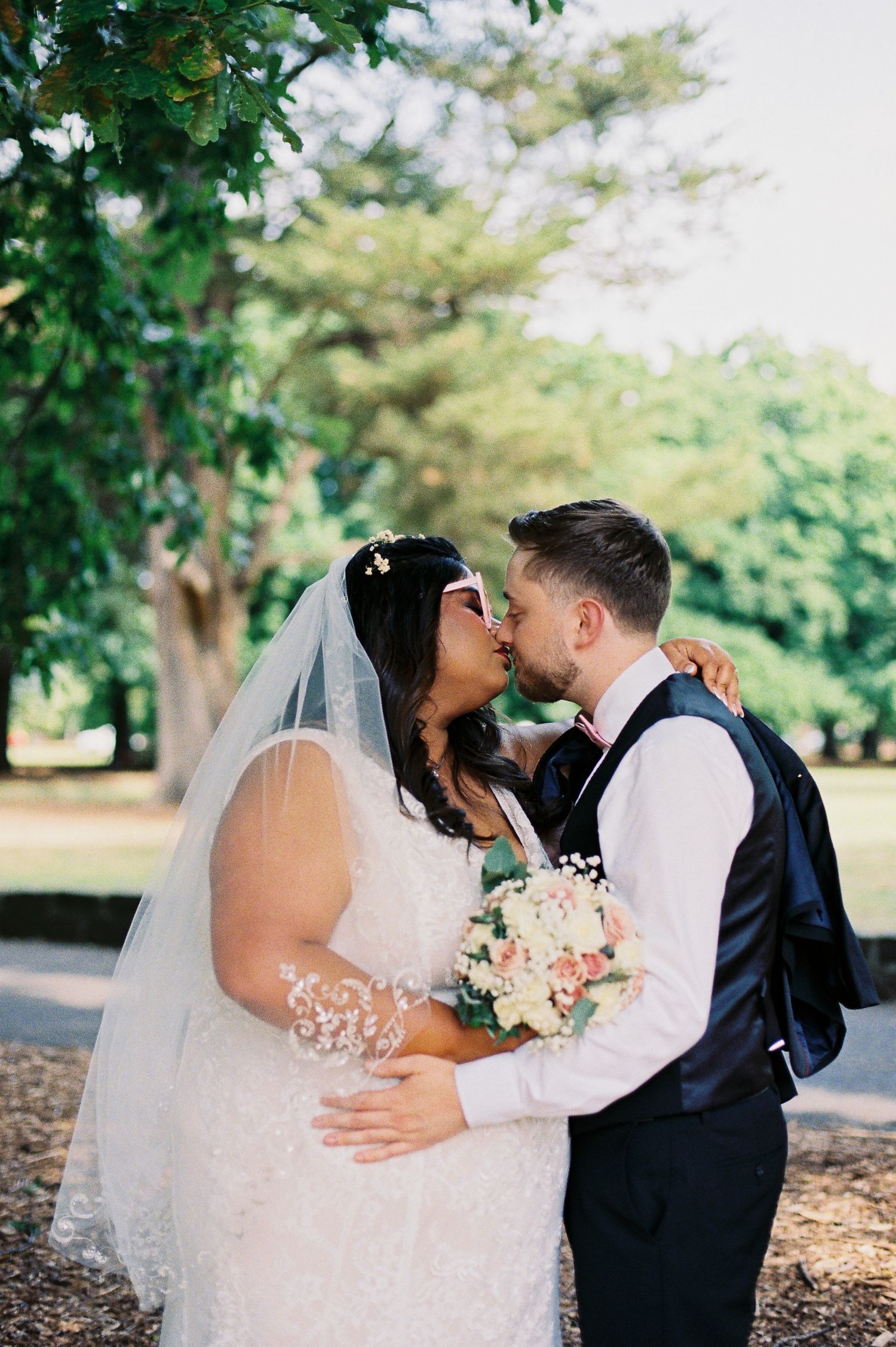 Bride and groom share a romantic kiss outdoors, bride in lace gown with veil holding a bouquet, groom in navy suit.