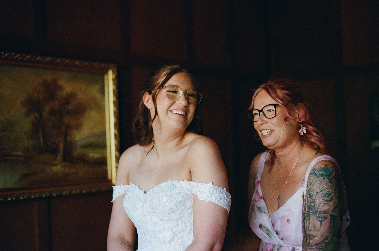 Bride in off-shoulder lace gown smiles with tattooed bridesmaid in floral dress, both wearing glasses.