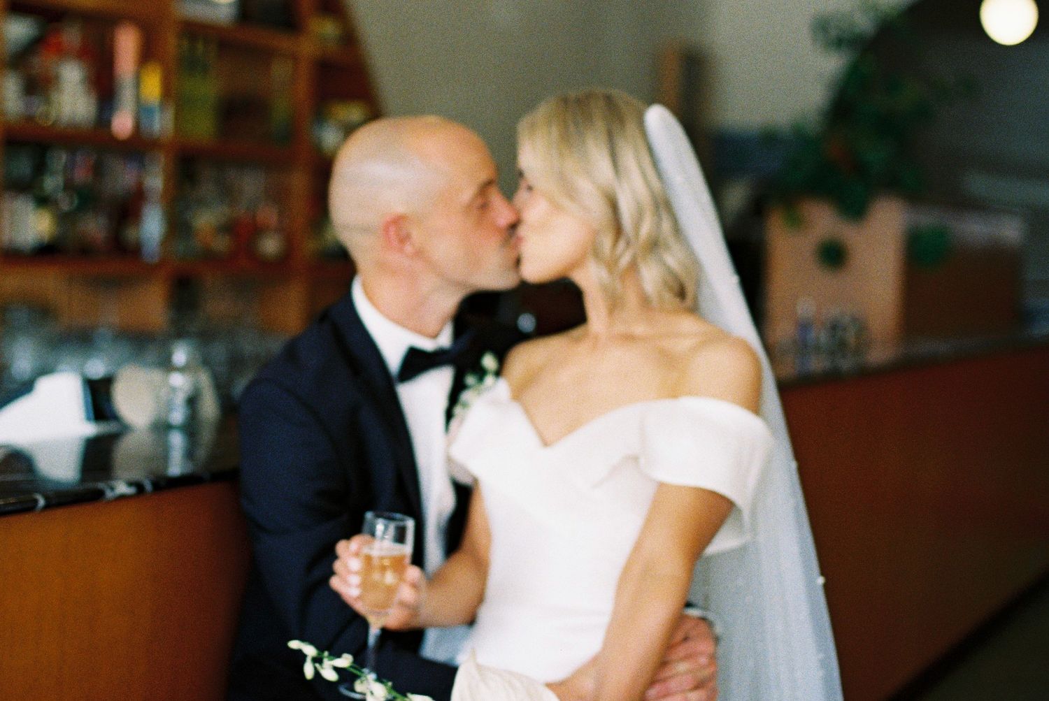 Bride and groom sharing a romantic kiss at a bar venue, bride in off-shoulder gown with veil, groom in navy tuxedo.