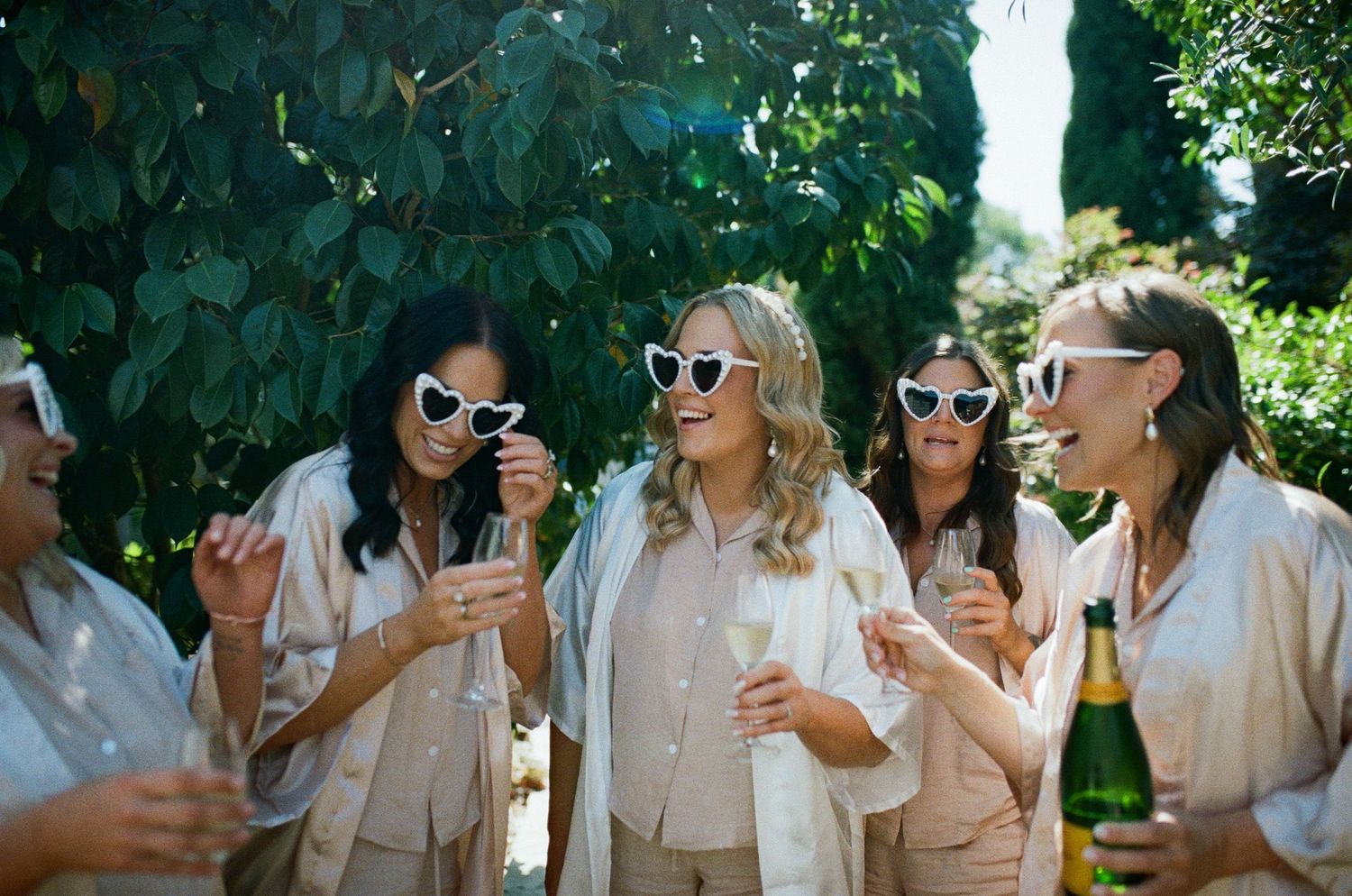 Bridesmaids in neutral satin robes wearing heart-shaped white sunglasses toasting with champagne outdoors.
