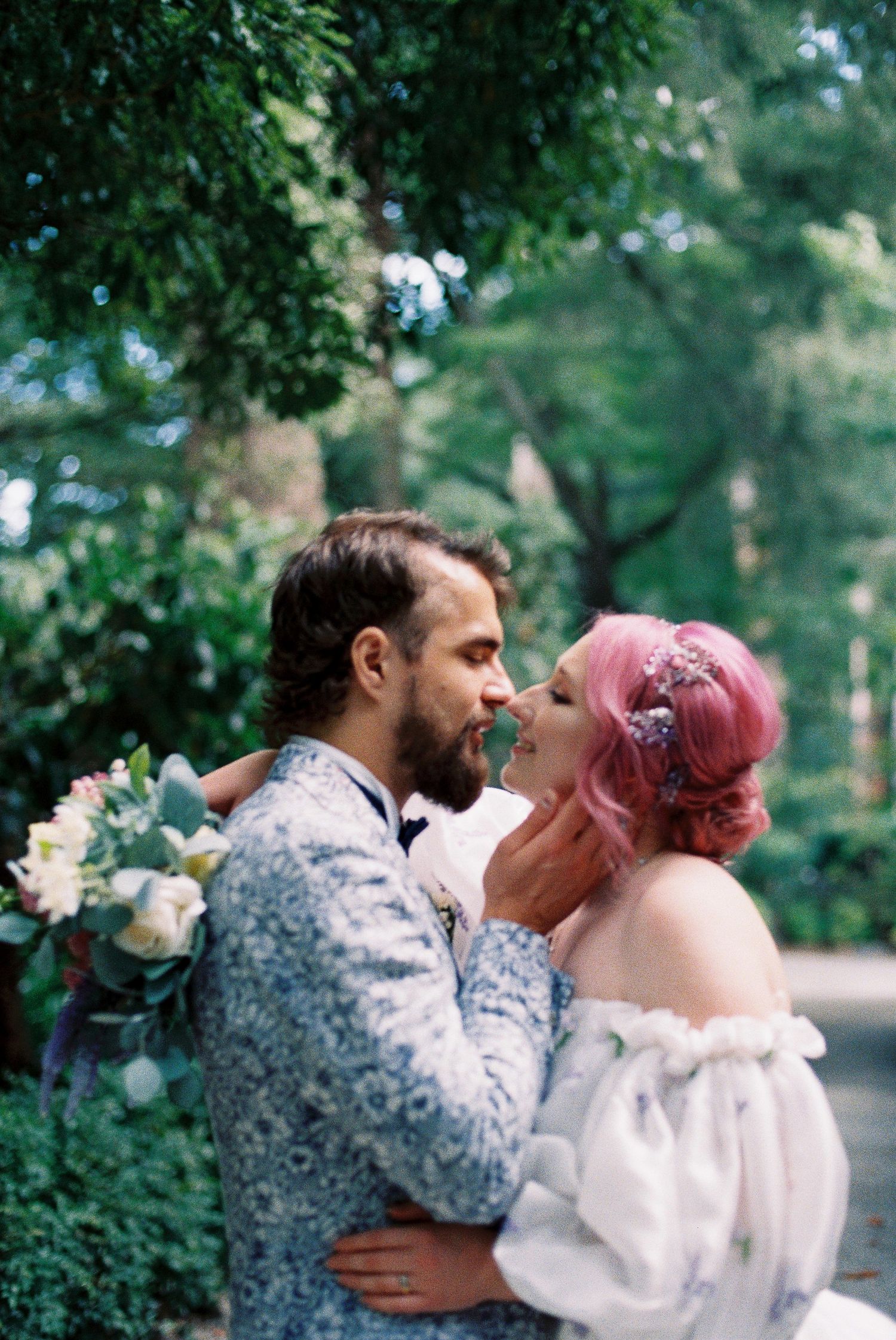 A bride with pink hair and groom in floral suit share a romantic moment outdoors surrounded by lush green trees.