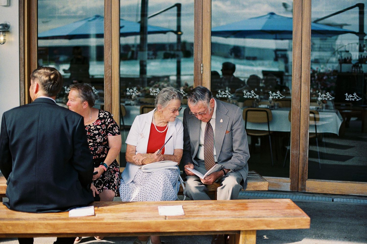 Elderly couple reading a booklet together at an outdoor cafe bench, with other guests seated nearby.