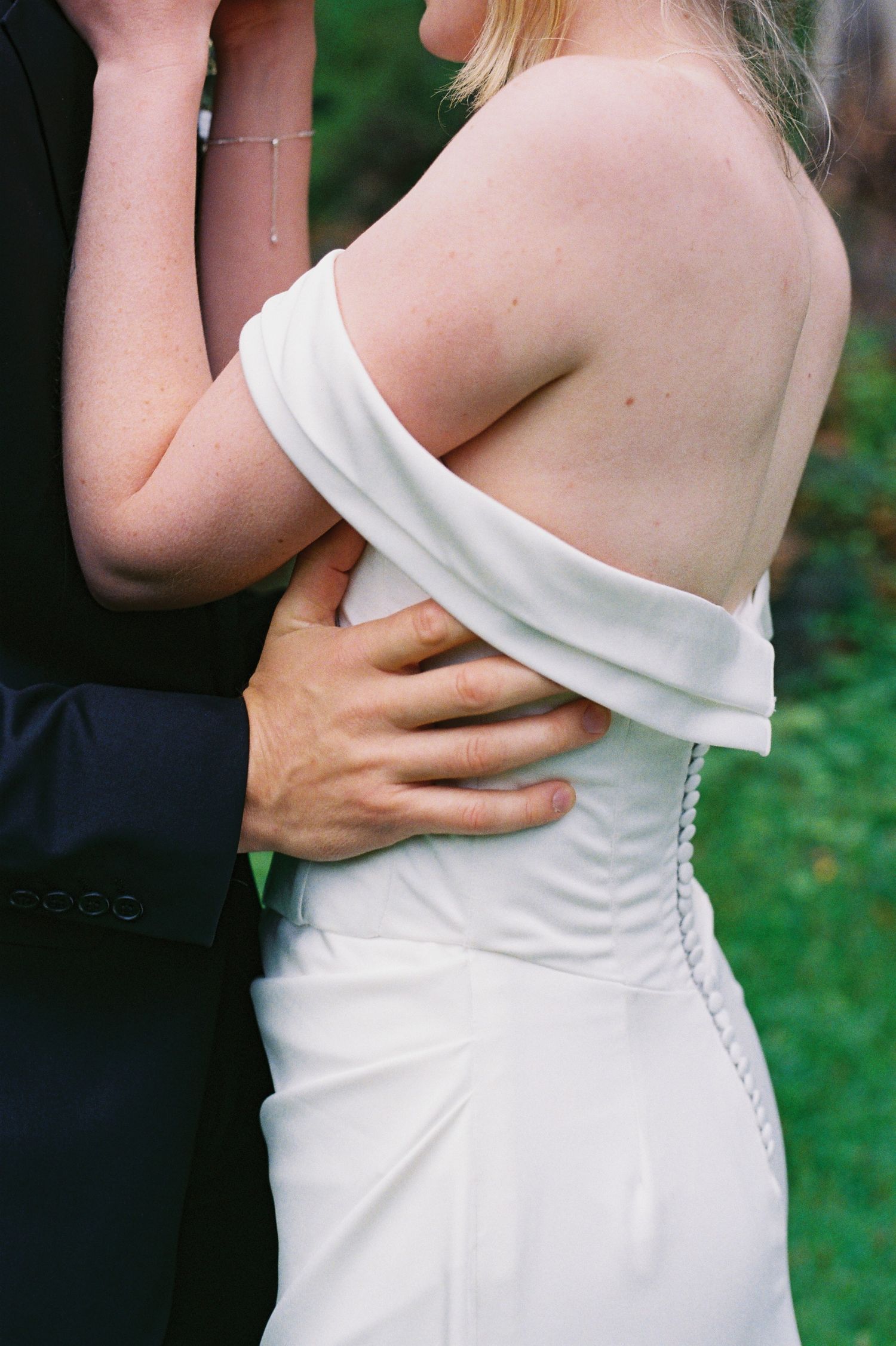 Bride in off-shoulder white wedding gown embraced from behind by groom in dark suit outdoors.