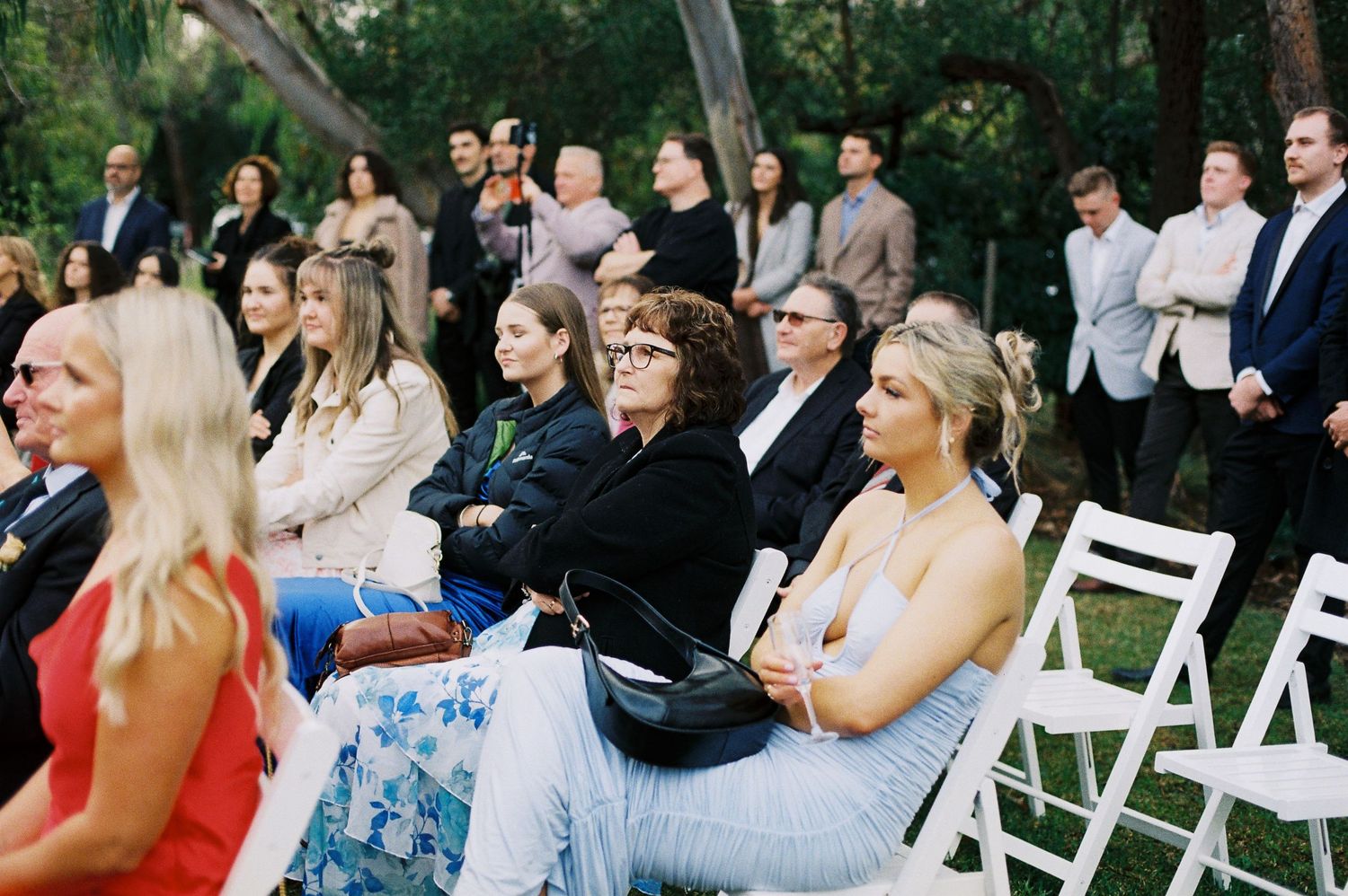Guests seated on white chairs at an outdoor wedding ceremony, dressed in elegant attire, enjoying the celebration.