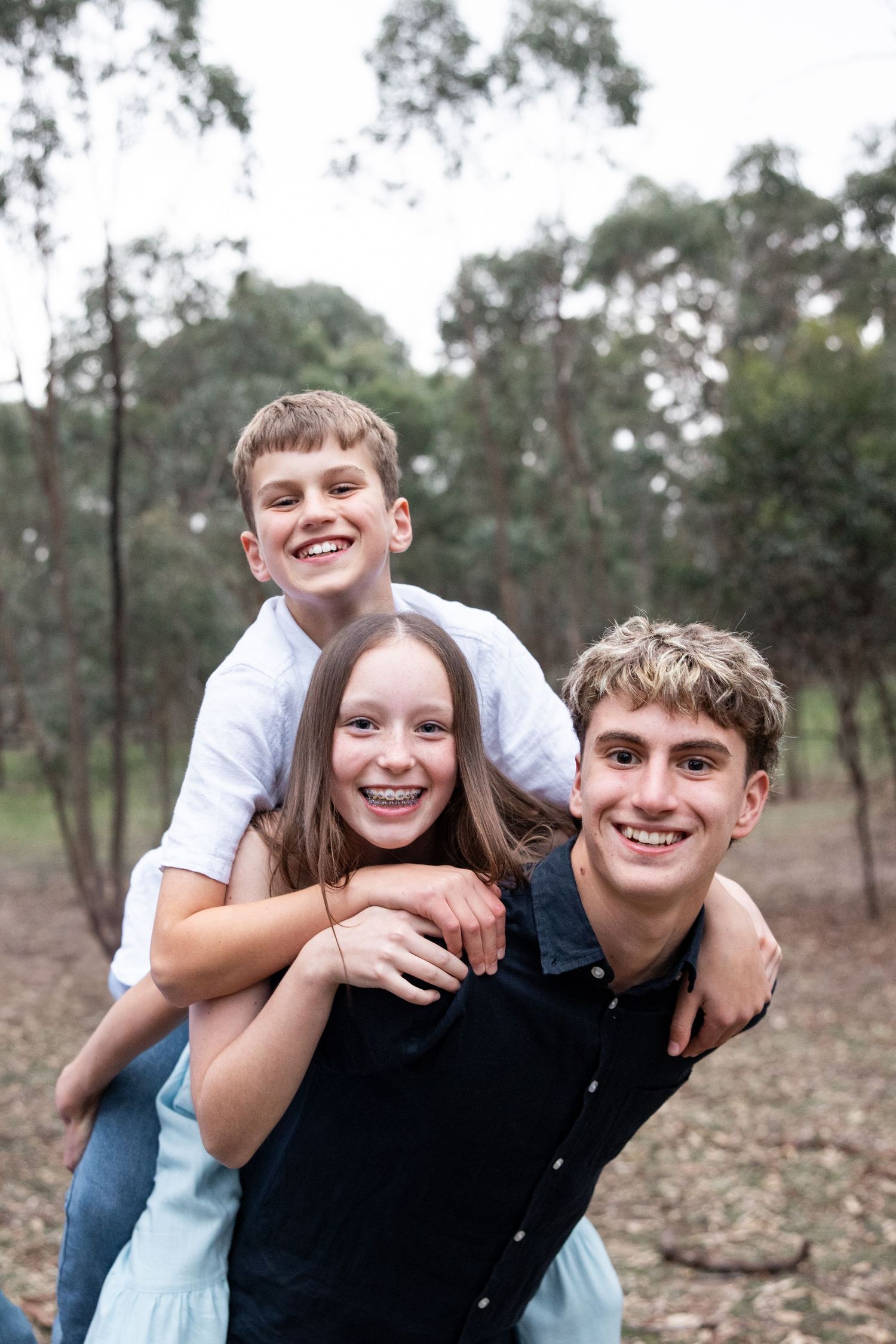 Three smiling teenagers pose together outdoors in a forest, with one giving a piggyback ride in a candid family photo.
