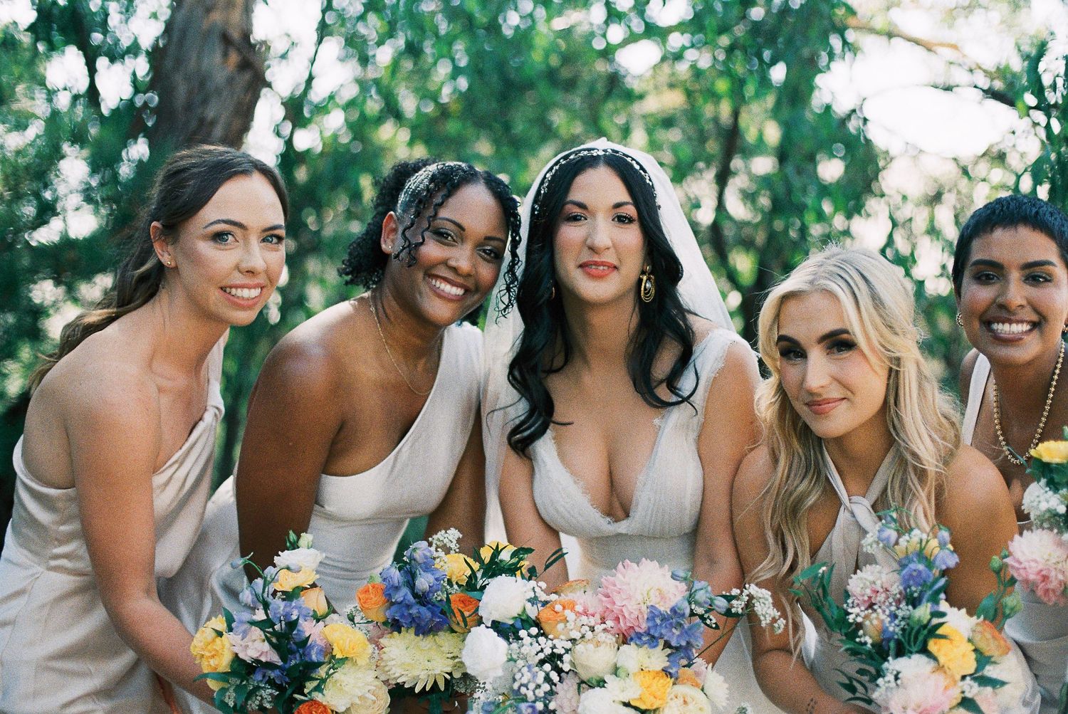 Bride in white gown poses with four bridesmaids in champagne dresses holding colorful floral bouquets outdoors.