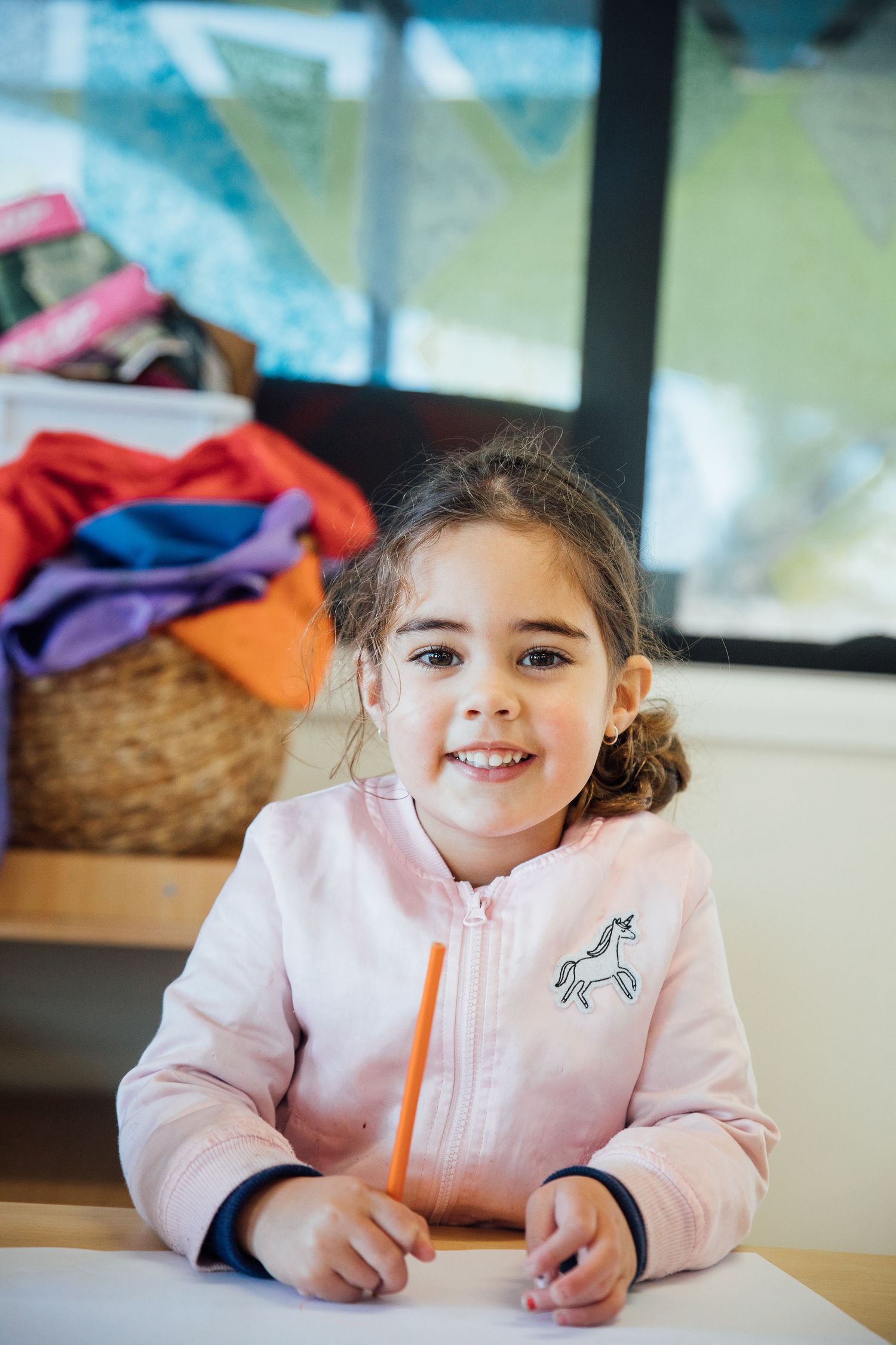 Child wearing pink zip-up sweater with unicorn design sits at desk with orange pencil doing schoolwork.