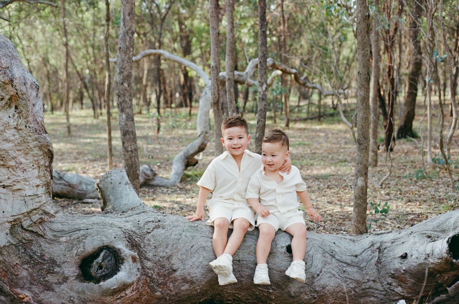 Two young boys in matching white outfits sitting on a large tree log in a natural woodland setting.