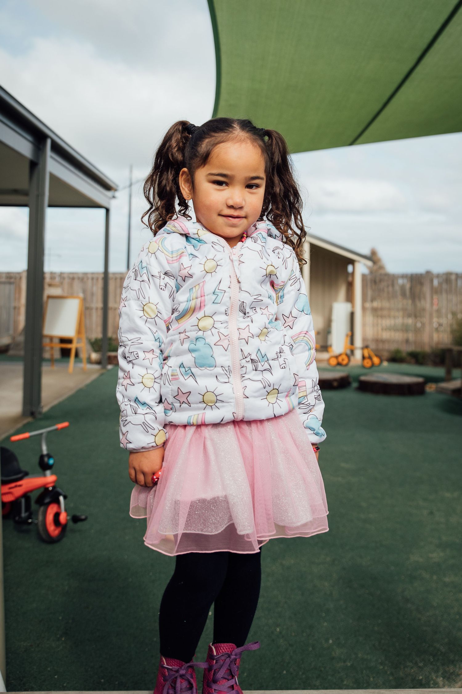 A child in a unicorn-print jacket and pink tulle skirt plays on a playground under a green shade canopy.