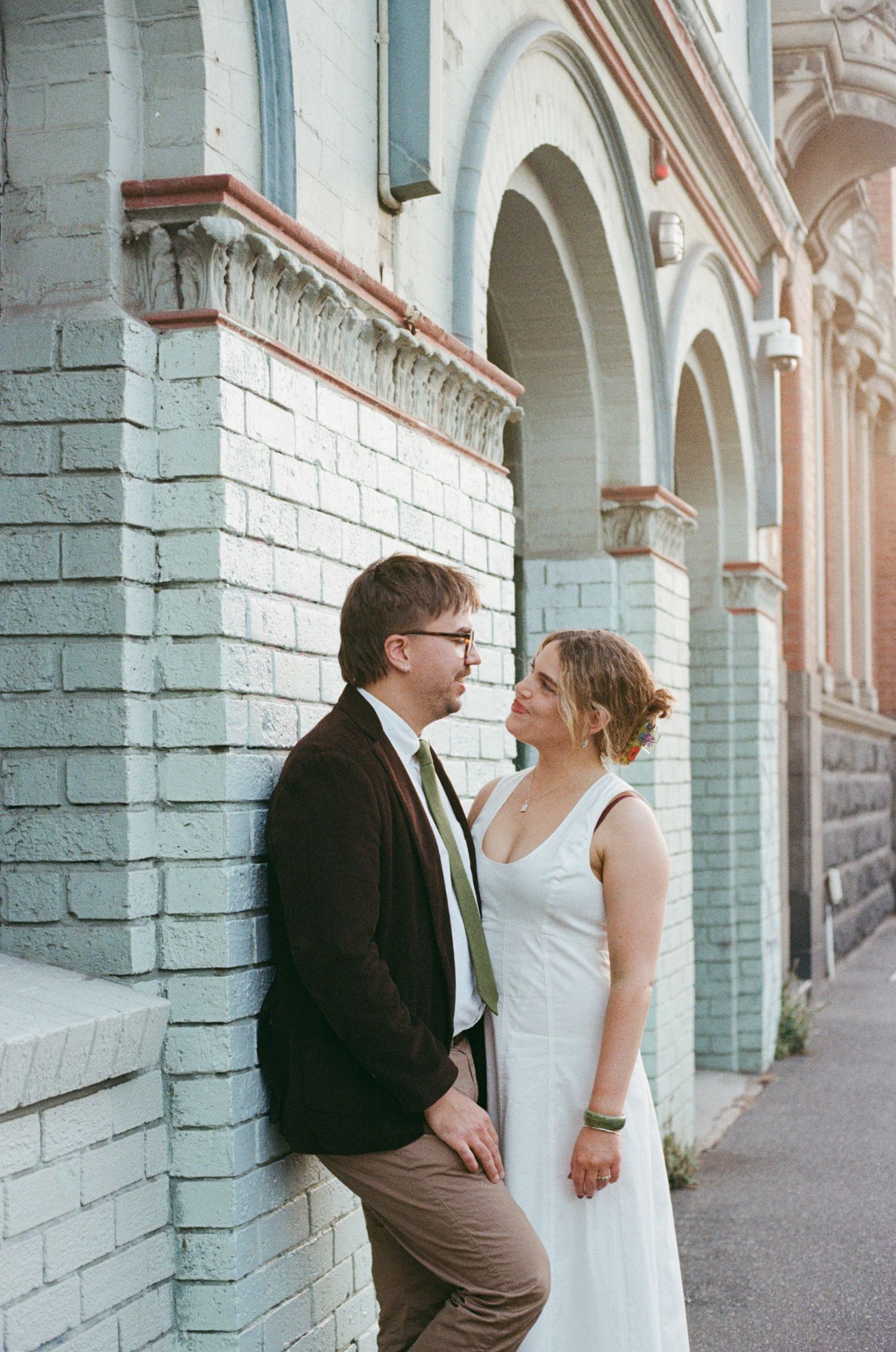 A couple in wedding attire share a romantic moment leaning against a light blue brick building with arched details.