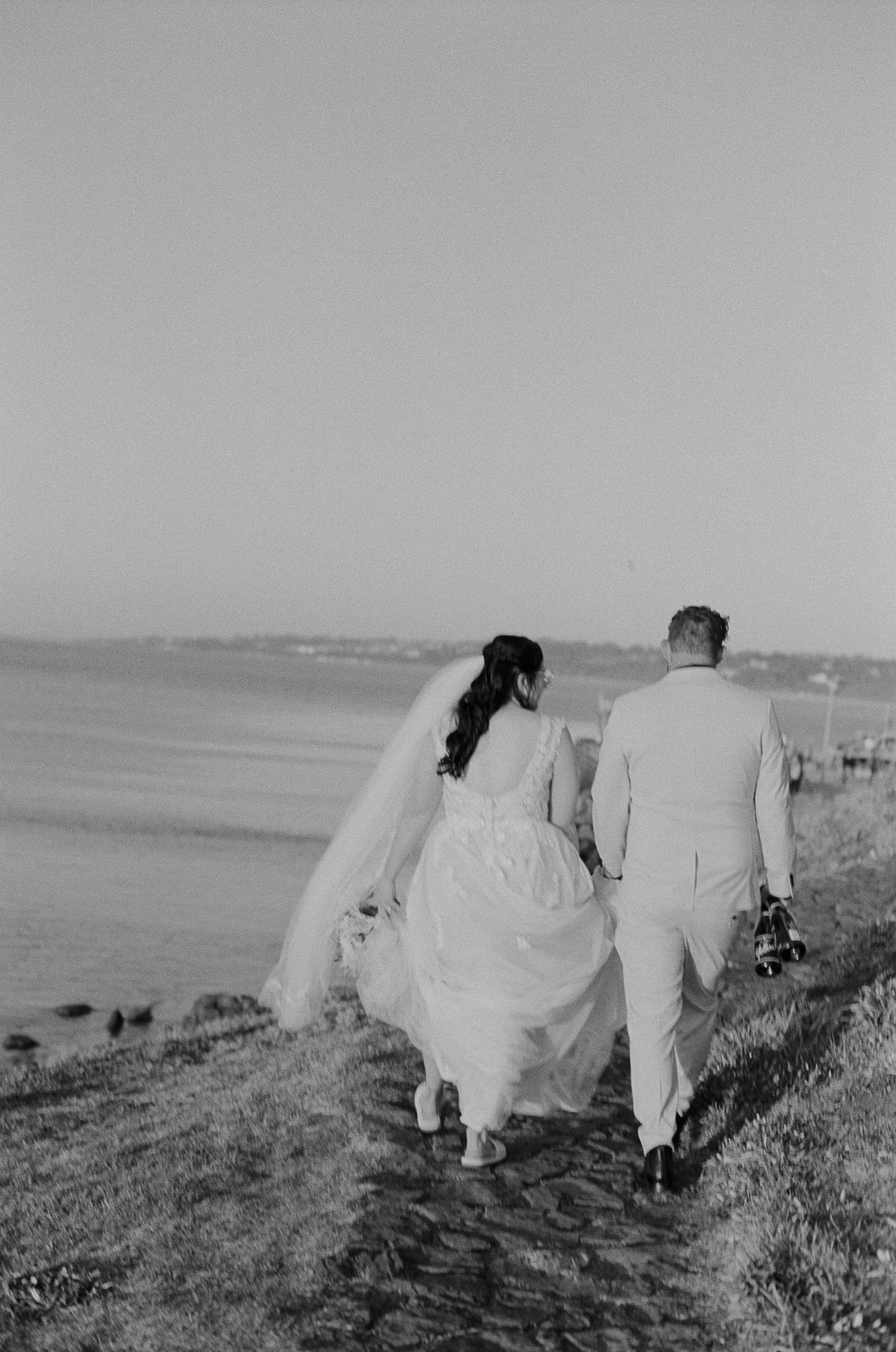 Black and white photo of a bride and groom walking hand in hand along a scenic waterfront path on their wedding day.