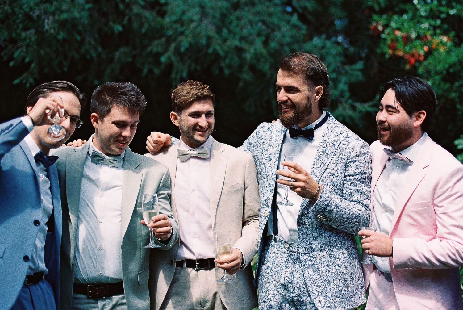 Five groomsmen in stylish suits celebrating outdoors, with the groom in a floral blue jacket holding a drink.
