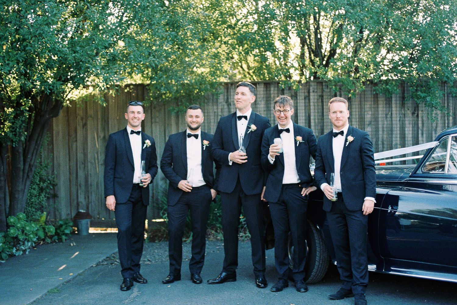 Five groomsmen in black tuxedos with bow ties pose outdoors near a classic black car before a wedding ceremony.