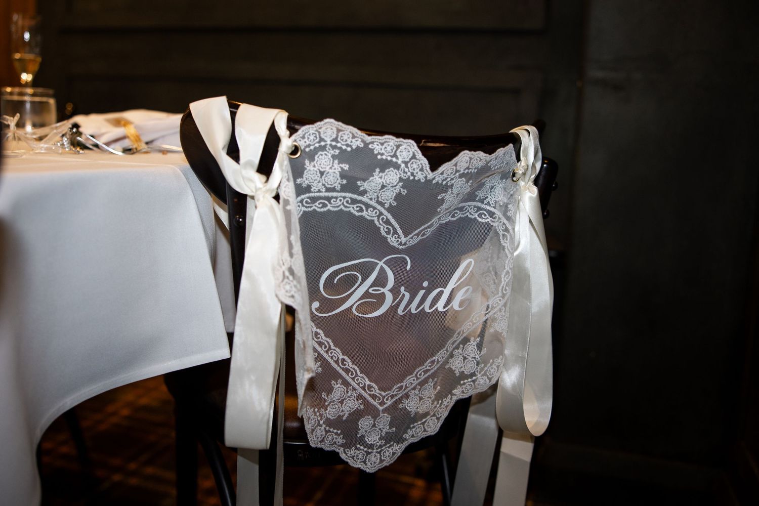 Elegant lace heart-shaped Bride chair sign tied with white satin ribbons on a dark wooden wedding reception chair.