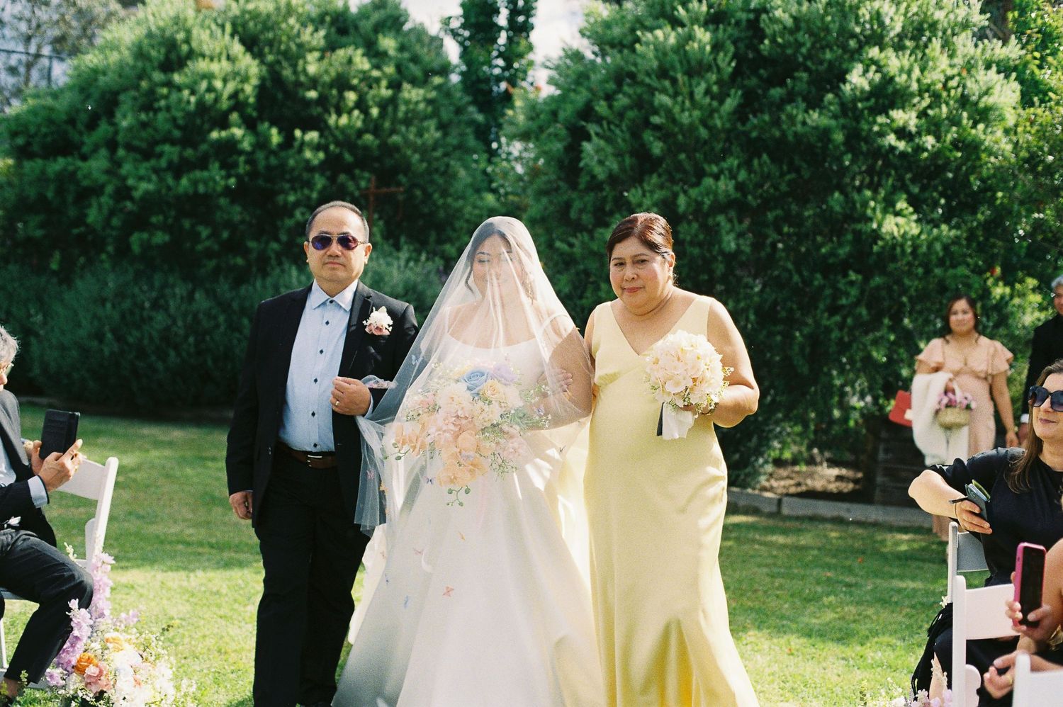 Bride in white gown and veil walks down outdoor aisle with parents at a sunny garden wedding ceremony.