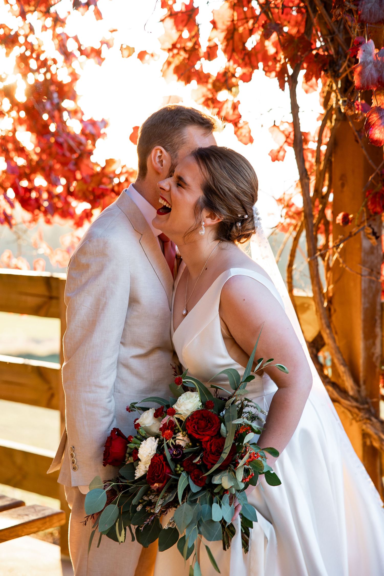 A romantic autumn wedding scene with white attired couple embracing against vibrant red fall foliage while holding a rustic bouquet.