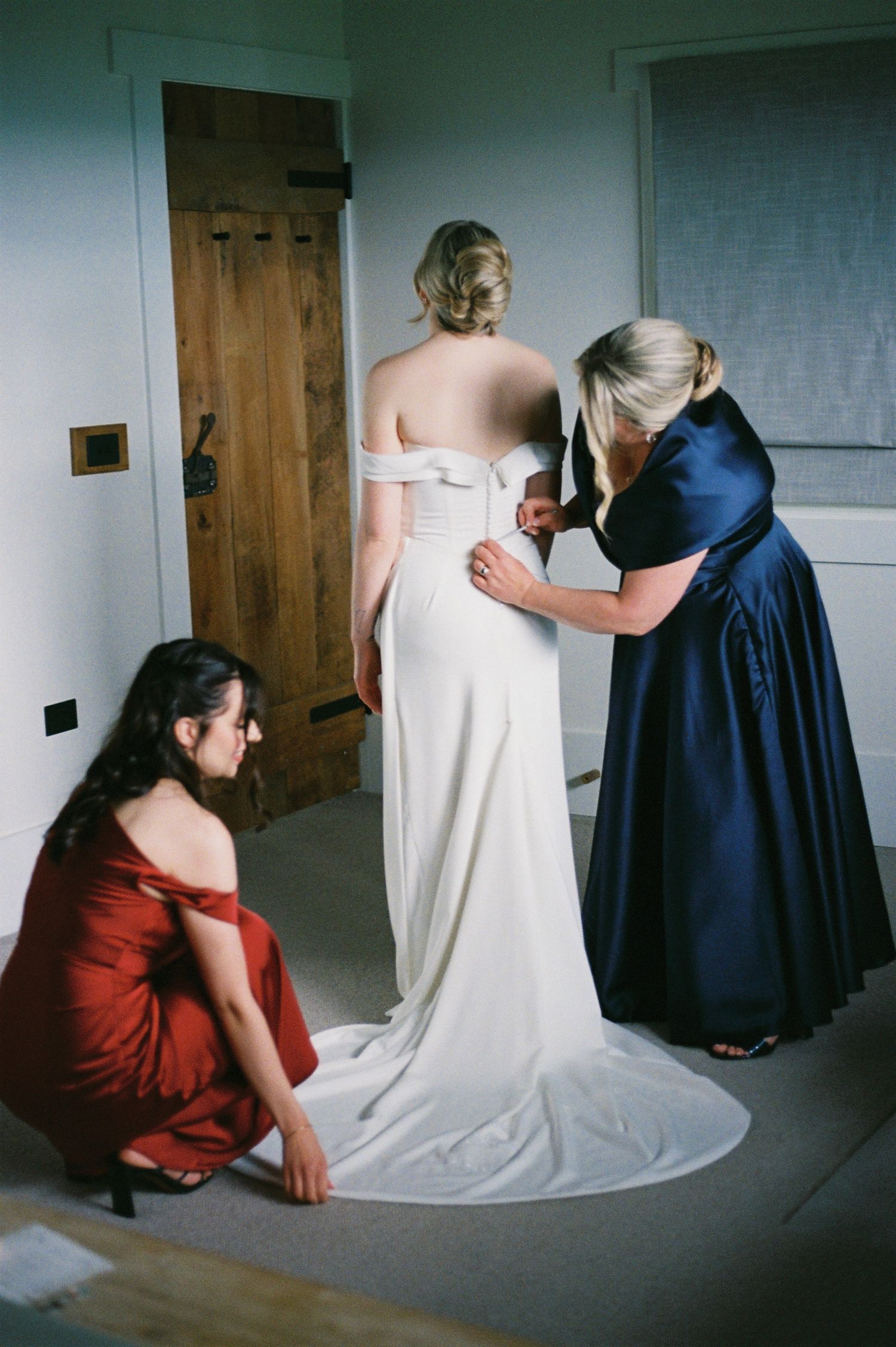 Bride in white off-shoulder gown being dressed by two women, one in navy blue and one in red, in a bright room.