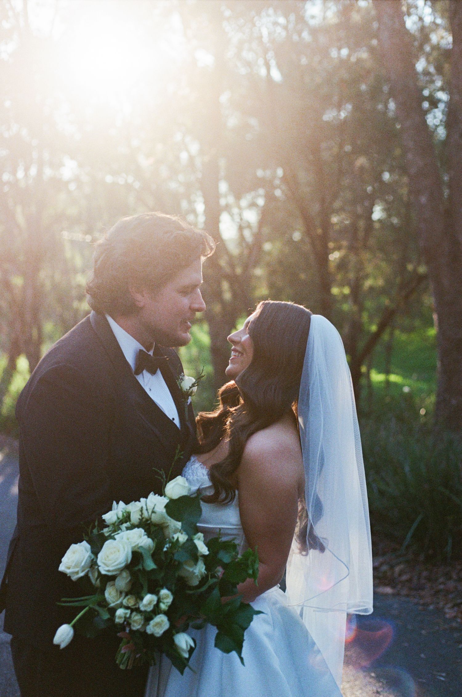 Bride and groom share a romantic moment in golden sunlight, bride holding white roses bouquet wearing veil.
