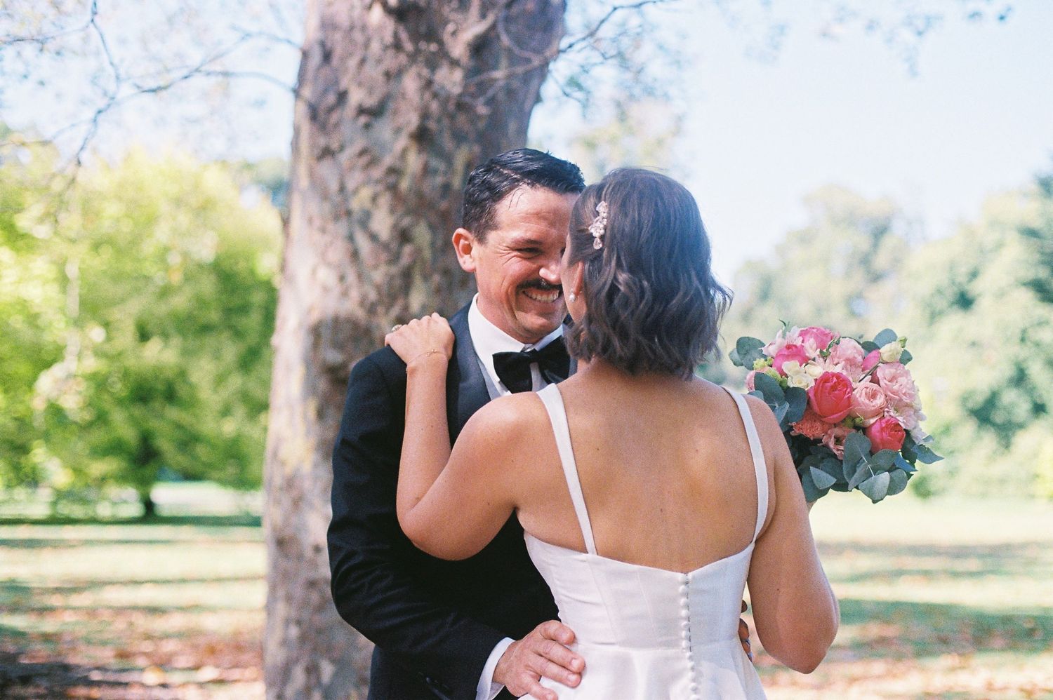 A bride and groom share a romantic moment outdoors, with the bride holding a pink bouquet and the groom in a black tuxedo.
