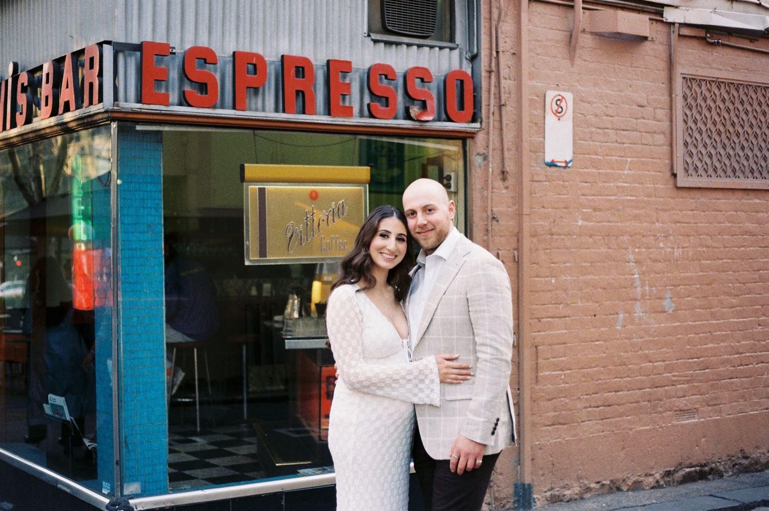 Couple embracing outside an espresso bar with neon signage on a brick wall street.
